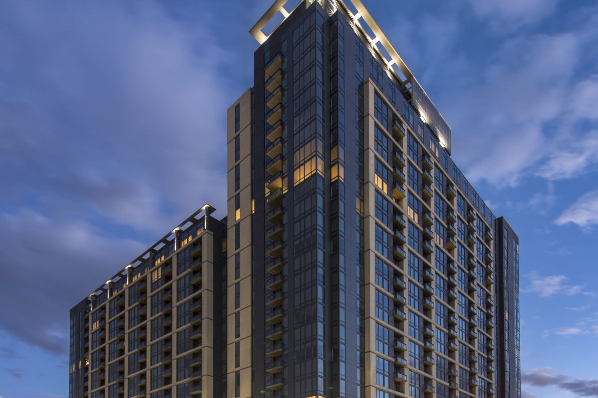 Modern high-rise building at dusk with illuminated windows and streetlights at a city intersection.