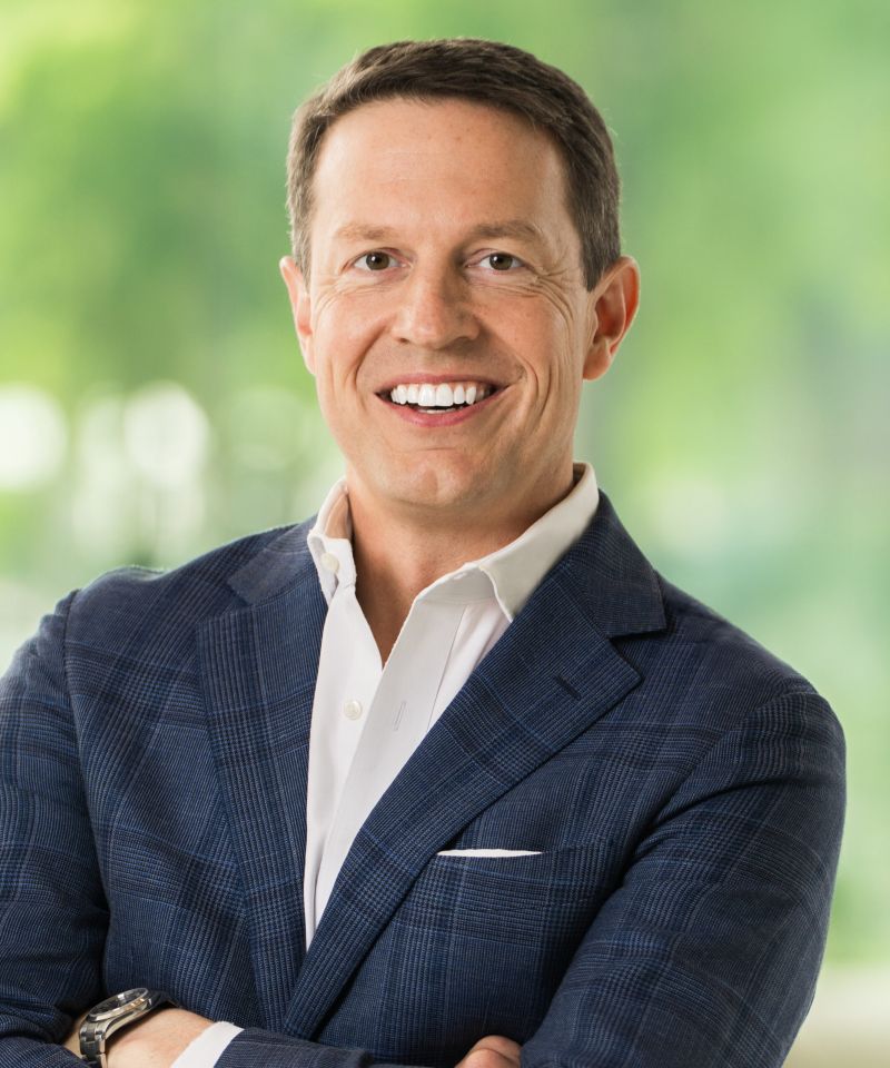 Smiling man in a blue checked suit jacket and white shirt, standing with arms crossed, blurred green background.
