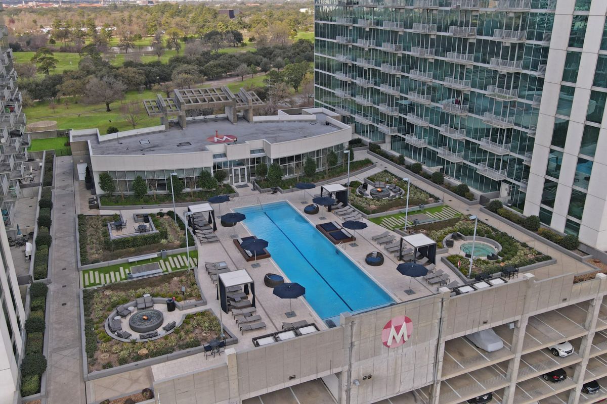 A rooftop pool area with lounge chairs, umbrellas, hot tub, and landscaping between two modern buildings.