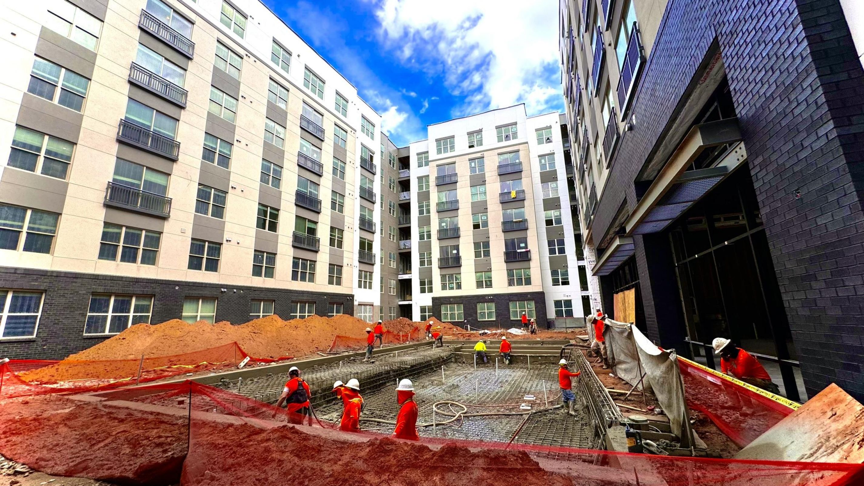 Construction workers in orange vests laying cement at a site surrounded by multi-story buildings under a blue sky at a Hanover Corporate construction project.