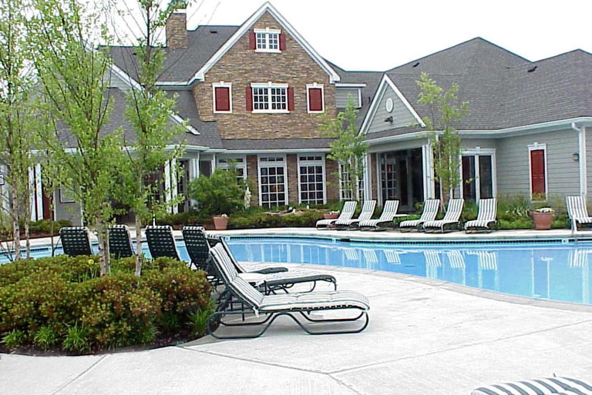 Outdoor swimming pool with lounge chairs in front of a large house with red shutters and landscaped greenery.