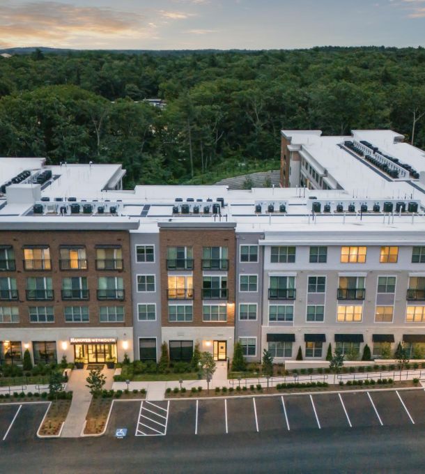 Aerial view of a modern apartment building with surrounding parking and forest in the background at sunset.