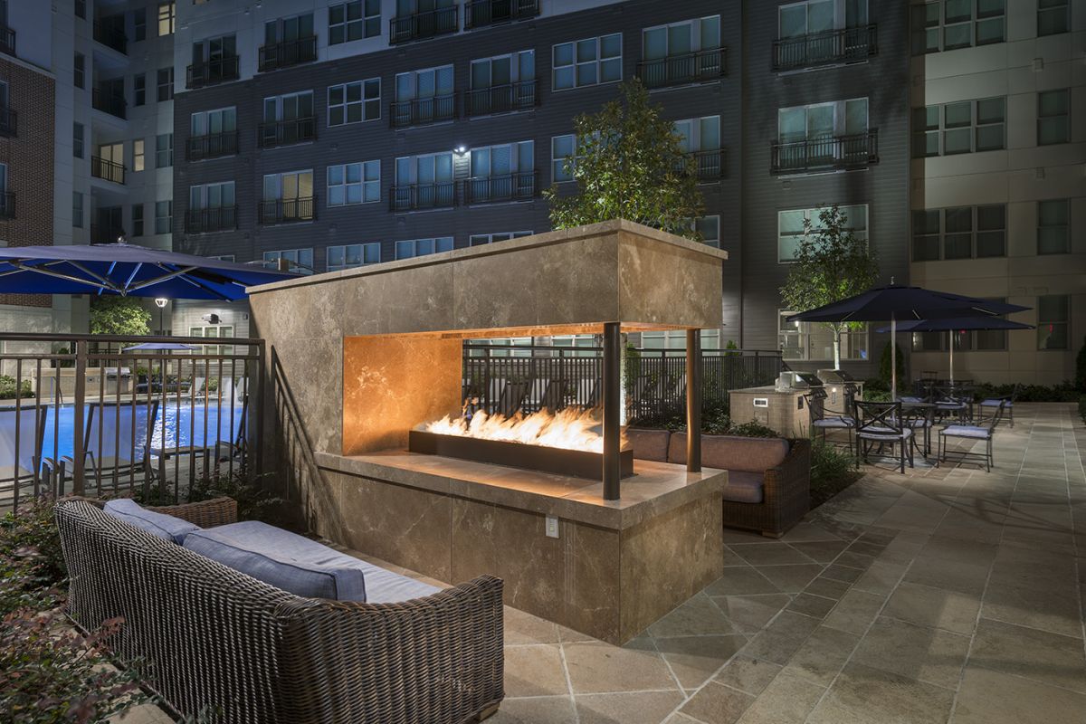 Outdoor patio at night with a modern fire pit, wicker seating, and poolside lounge area.
