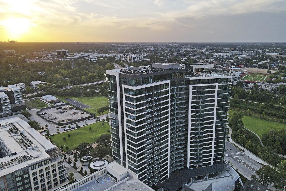 Aerial view of a tall, modern building in a cityscape at sunset, surrounded by greenery and urban structures.