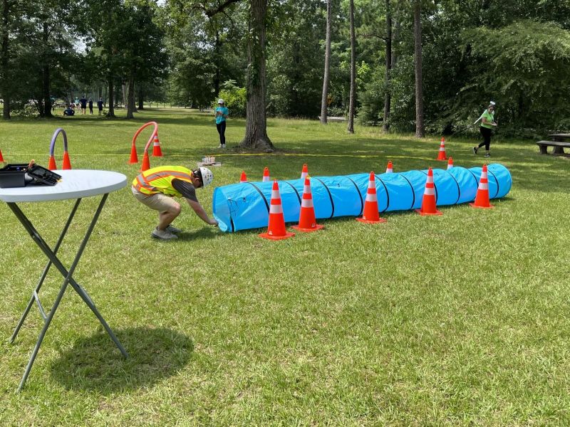 Person in a helmet and vest crawls through a blue tunnel surrounded by orange cones on a grassy field.