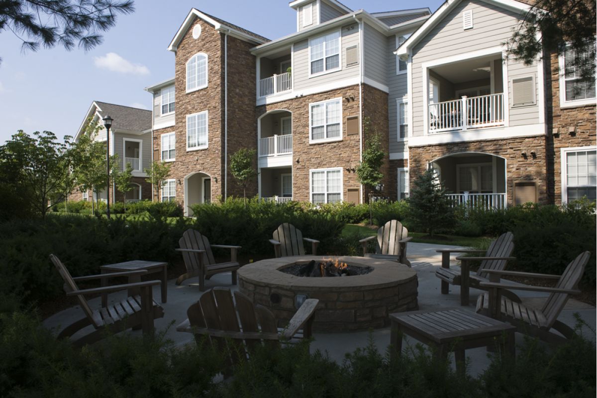 Outdoor fire pit with wooden chairs surrounded by landscaping in front of a modern apartment building.