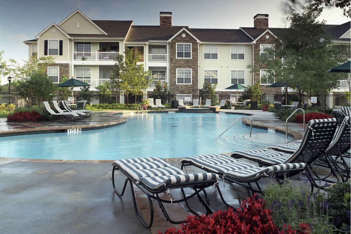 Outdoor pool with striped lounge chairs, surrounded by landscaping and an apartment building in the background.