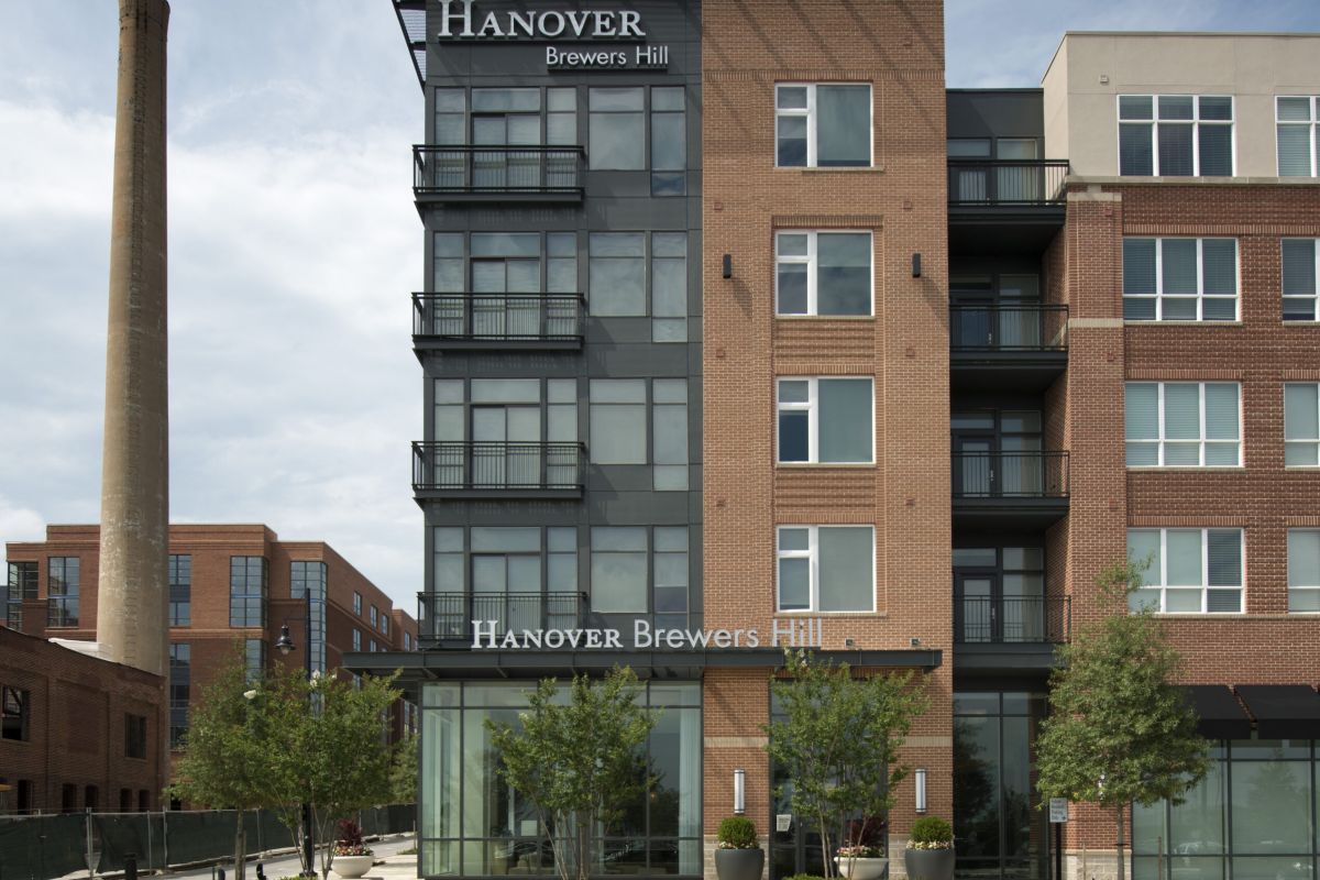 Modern apartment building with “Hanover Brewers Hill” signage, large windows, and a tall brick smokestack nearby.
