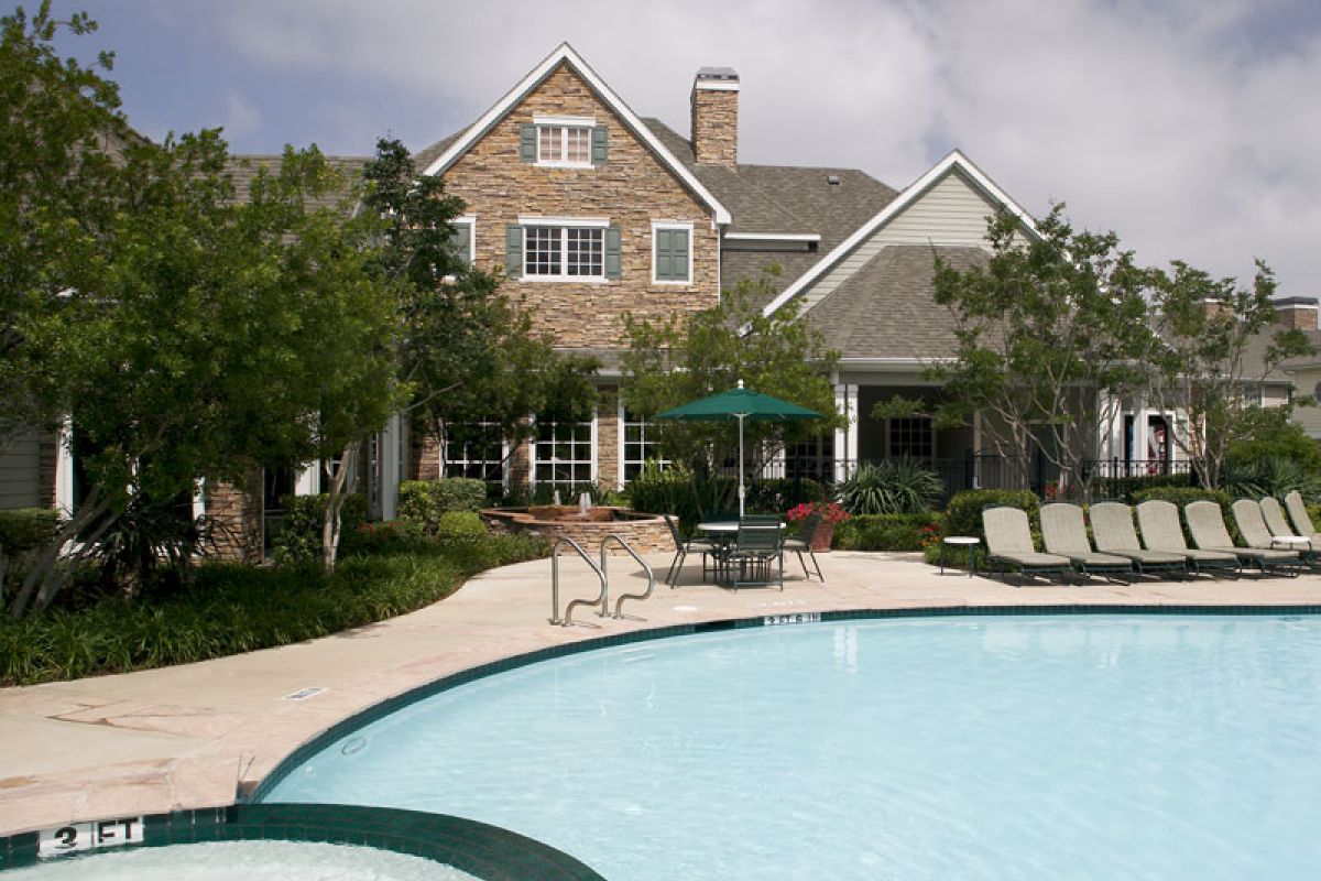 A large house with a stone facade behind a swimming pool and lounge chairs under a partly cloudy sky.