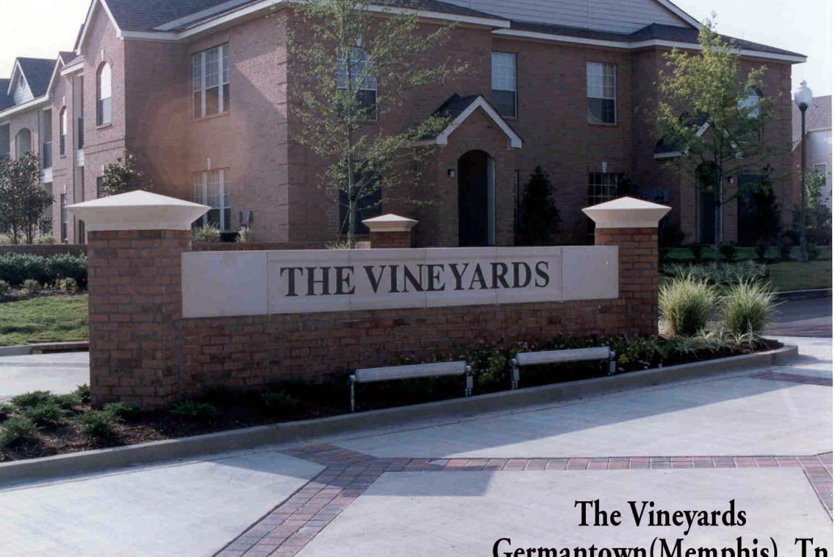 Brick sign reading "The Vineyards" at the entrance of a residential complex with townhouses in the background.