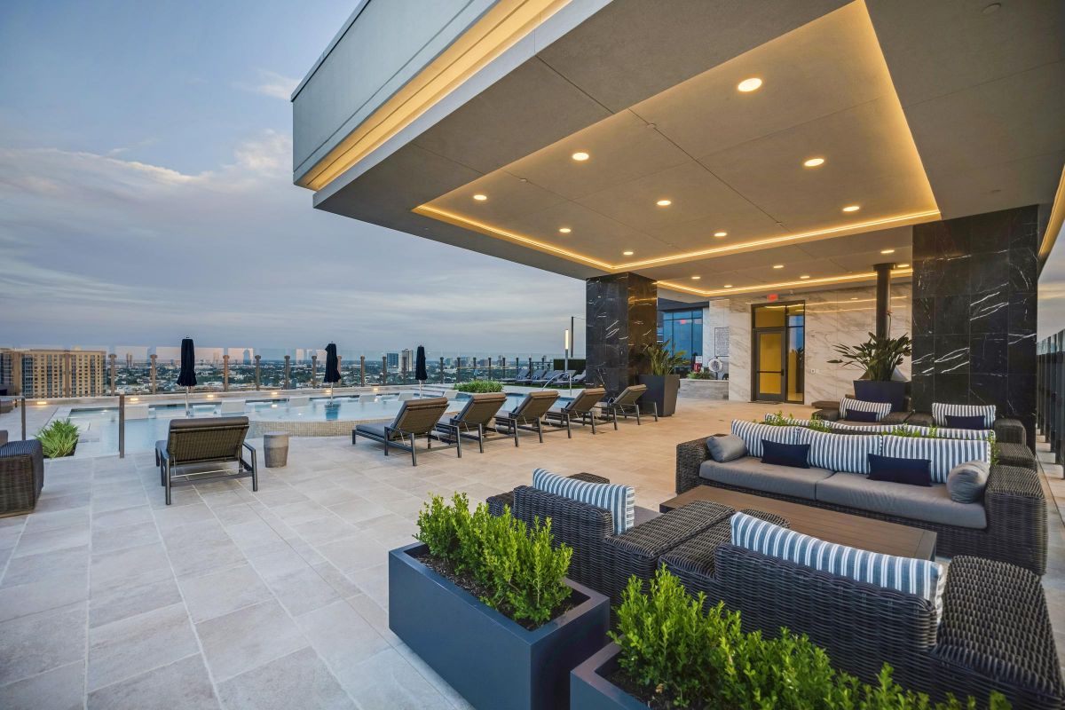 Modern rooftop lounge with sofas, lounge chairs, and a pool. Cityscape visible in the background under a cloudy sky.