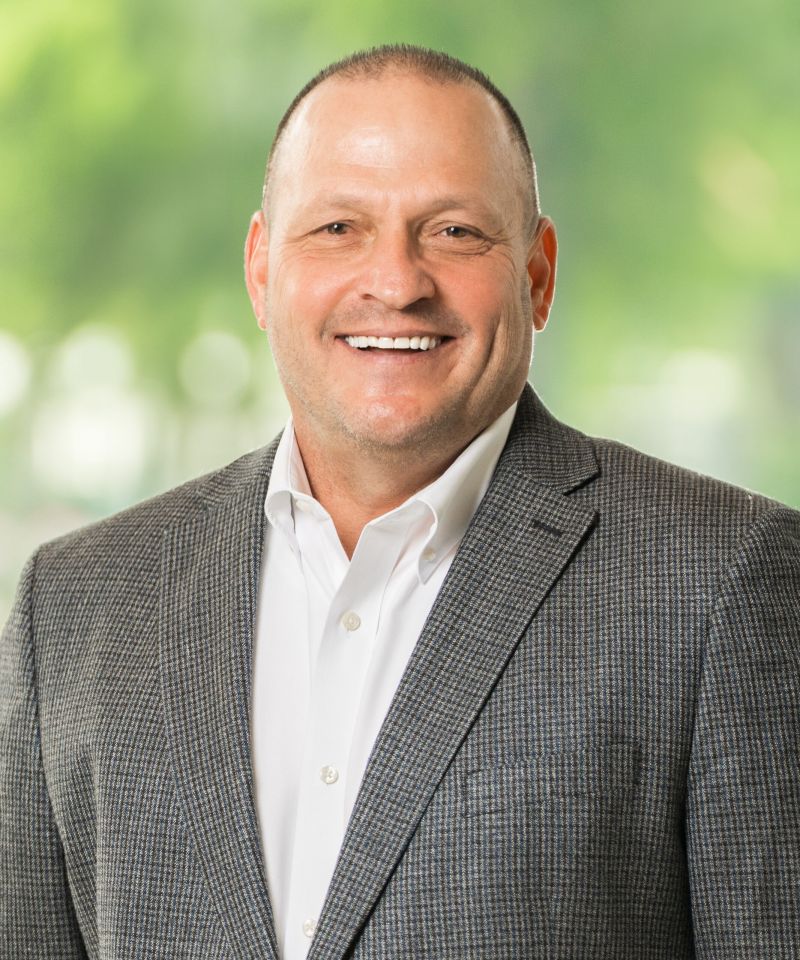Smiling man in a gray checked blazer and white shirt standing outdoors with a blurred green background.