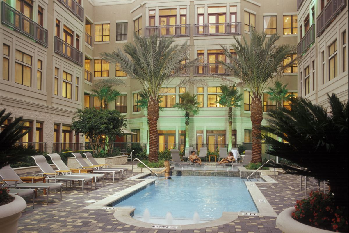 Courtyard pool with lounge chairs, palm trees, and people relaxing at a modern multi-story apartment building.
