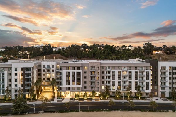 Modern apartment complex at sunset with a backdrop of trees and a colorful sky.