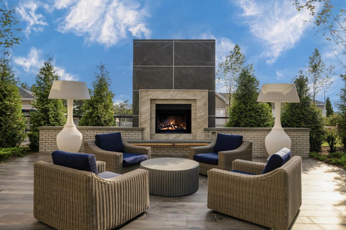 Outdoor patio with wicker chairs, a round table, two large lamps, and a central fireplace under a blue sky.