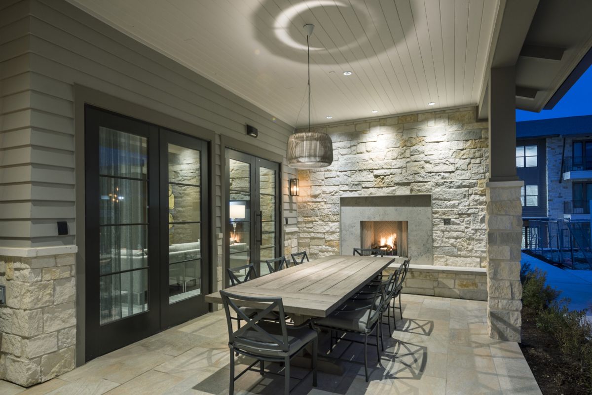Covered patio with a dining table, chairs, stone fireplace, and modern lighting at dusk.