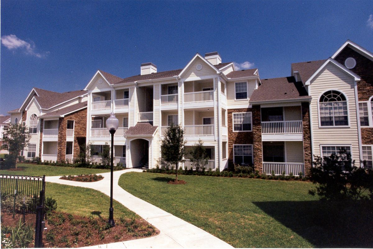 Three-story apartment building with balconies, brick and siding exterior, and landscaped lawn under blue sky.