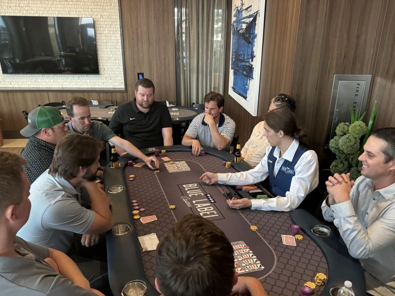 Eight men and a dealer play poker at a casino table, chips and drinks on the table in a modern room.