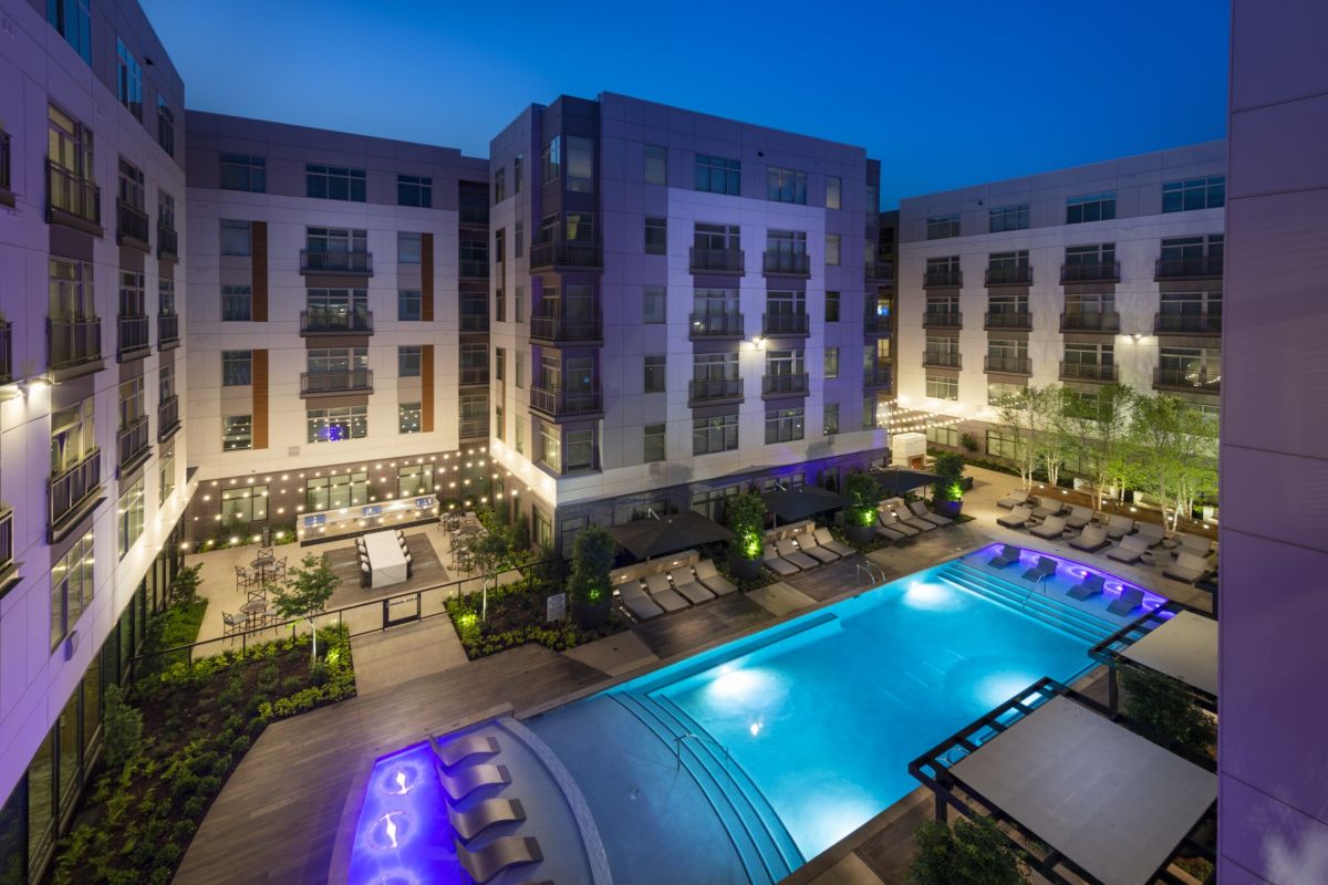 Modern apartment complex courtyard at dusk with illuminated swimming pool and lounge areas.