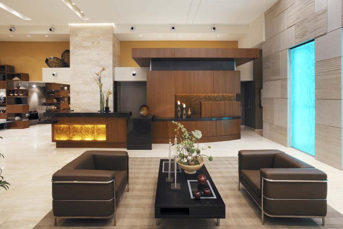 Modern hotel lobby with brown chairs, a coffee table, wood accents, and blue-lit water feature on the right wall.