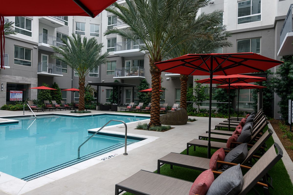 Modern Hanover Warner Center apartment pool area with lounge chairs, red umbrellas, palm trees, and surrounding balconies.