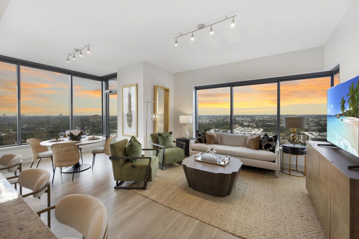 Modern living room with large windows at sunset, featuring cozy seating, a TV, dining area, and city views.
