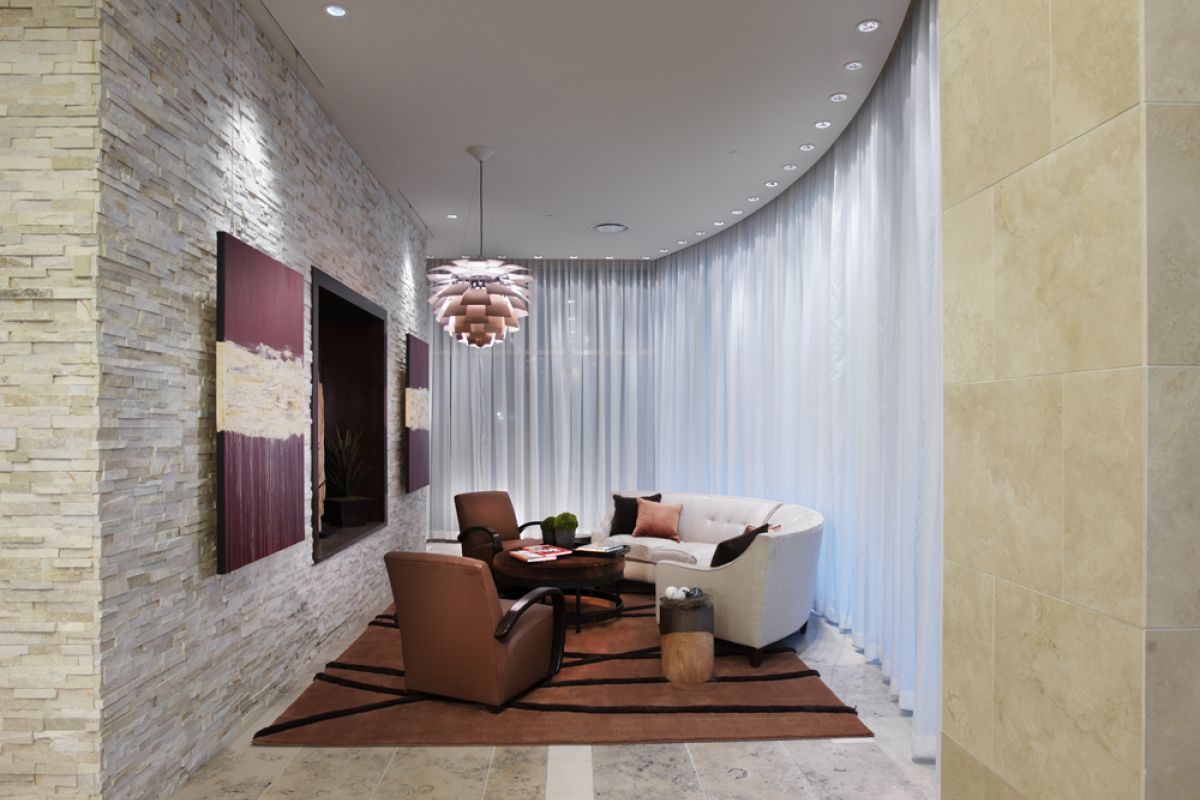 Modern lounge with armchairs, a sofa, abstract art, stone walls, and sheer curtains, lit by a pendant lamp.