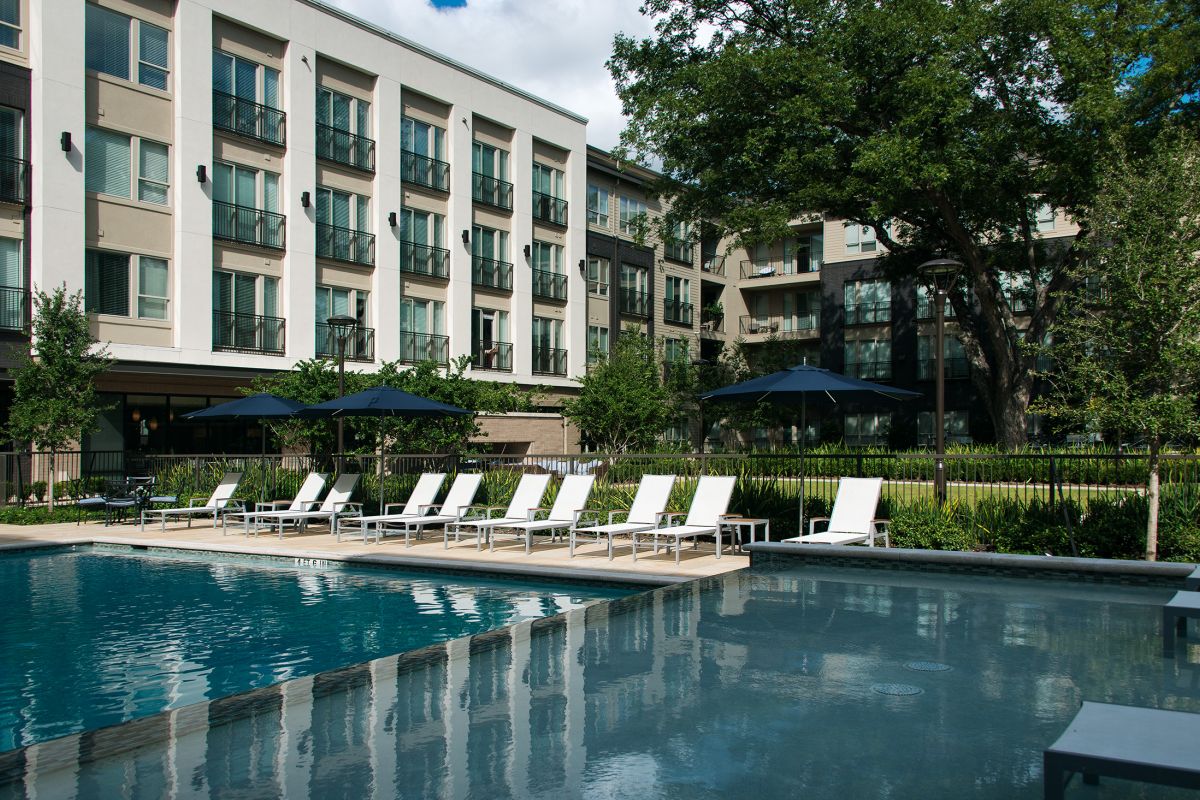 Modern apartment complex with a swimming pool, lounge chairs, and umbrellas, surrounded by greenery.