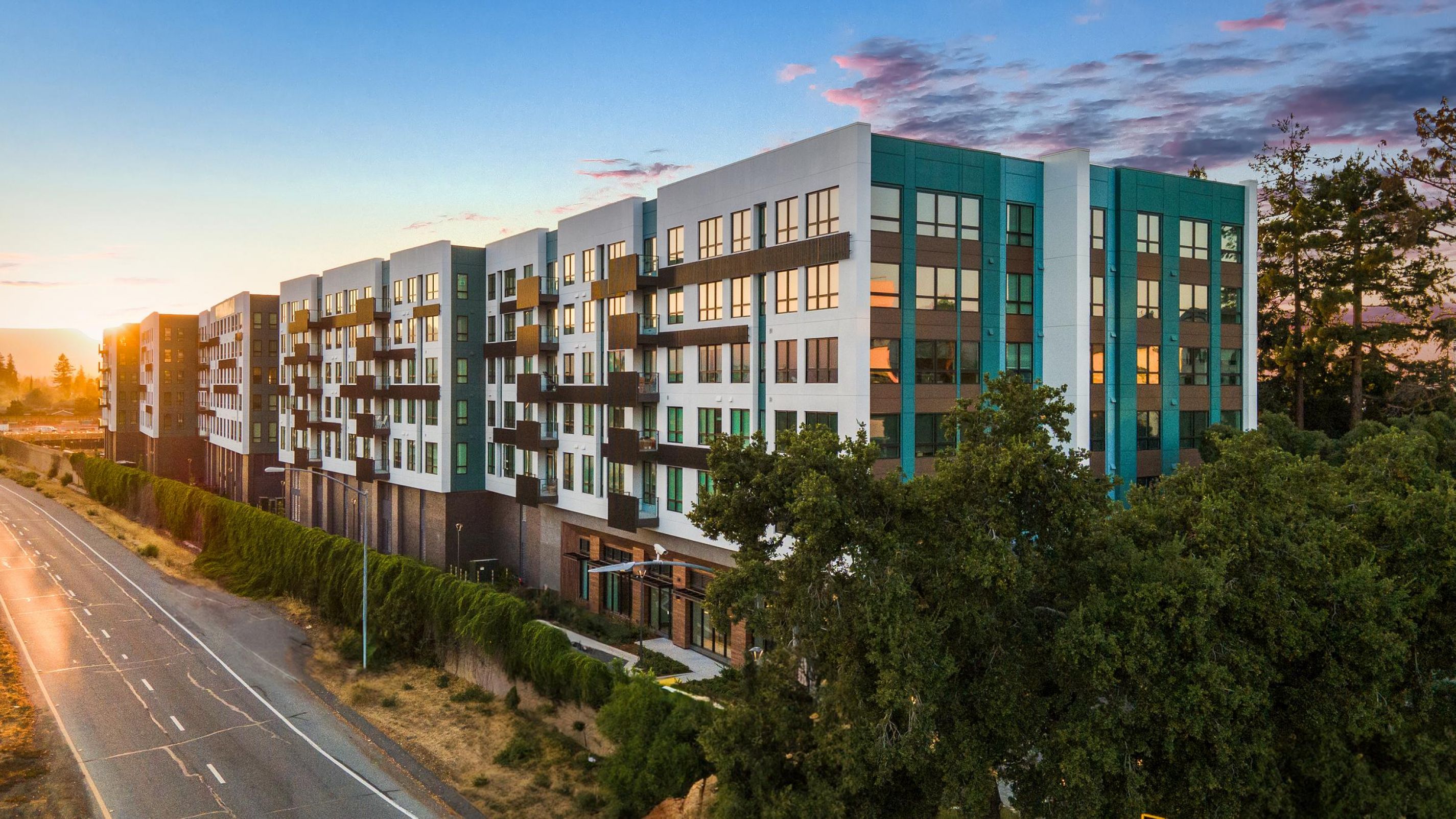 Modern apartment complex with large windows and balconies at sunset, adjacent to a road and surrounded by trees.