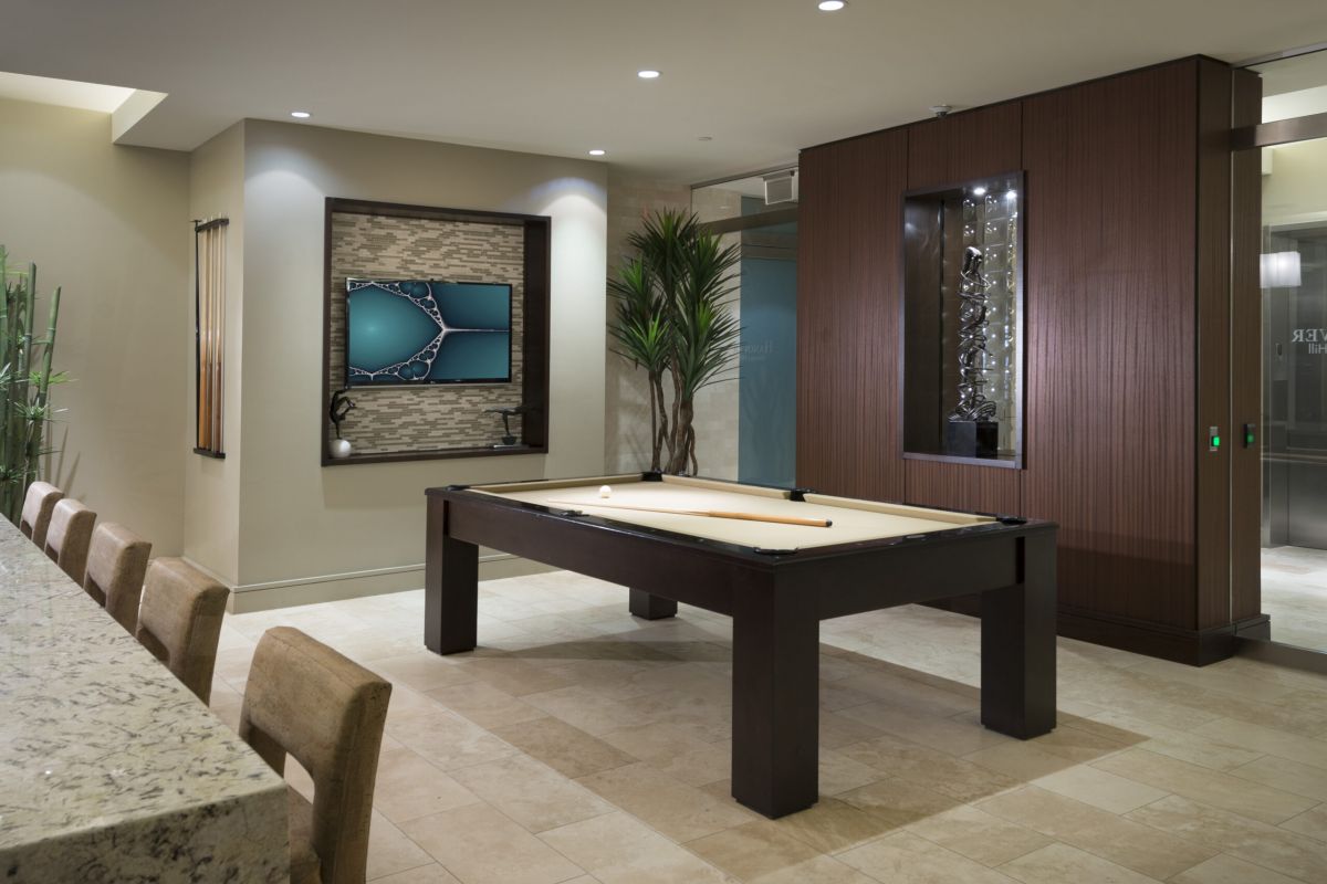 Modern game room with a pool table, wall art, countertop with chairs, and tall decorative plants.