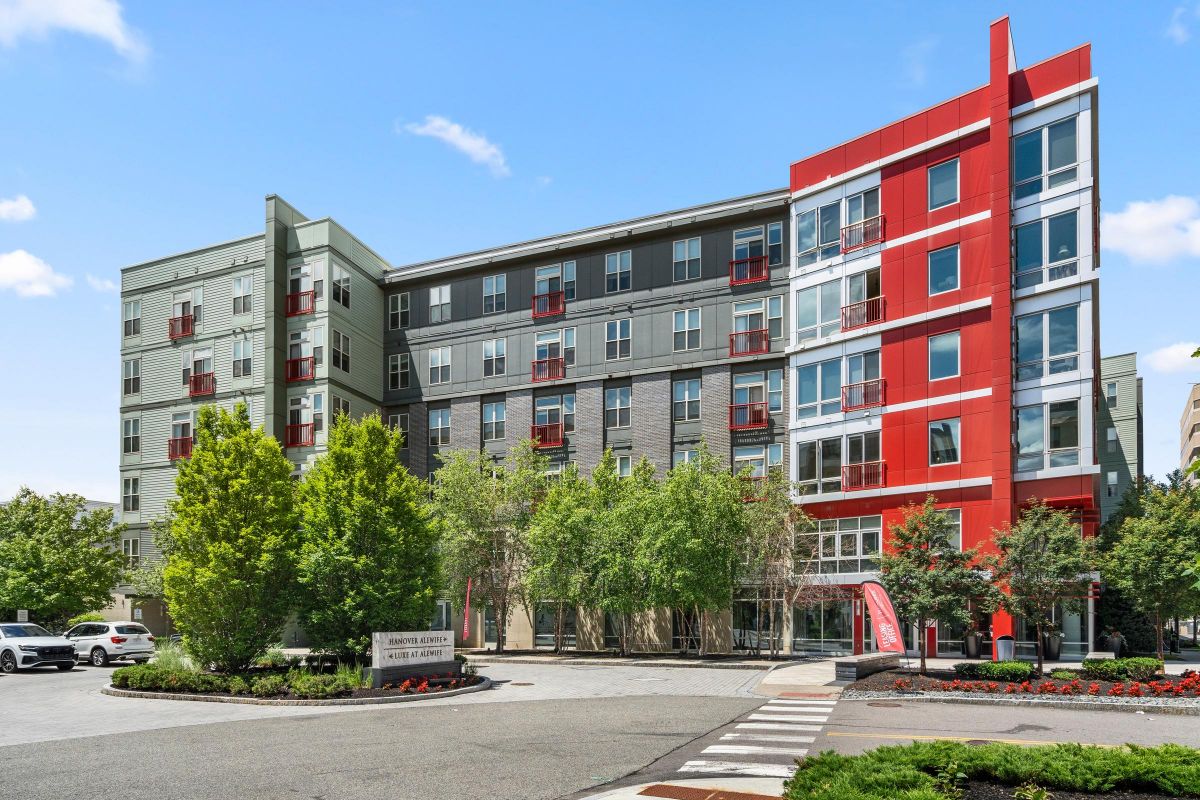 Modern multi-story apartment building with red and gray accents, surrounded by trees and a clear blue sky.